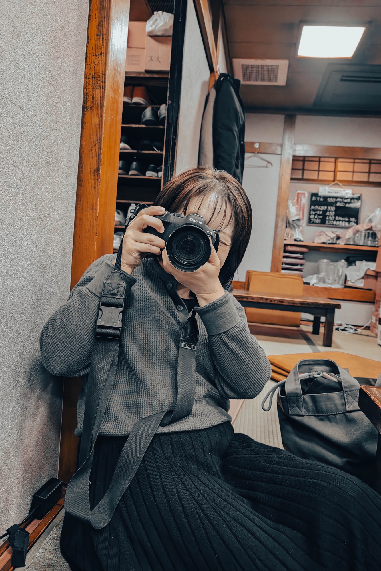 居酒屋の座敷で、キヤノンのミラーレスカメラを構えて満面の笑みを浮かべる女性のポートレート。