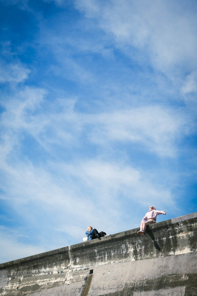 江ノ島の高い堤防に座り、両手を広げて青空を見上げる女性のシルエット。空が画面の大部分を占める開放的な構図。
