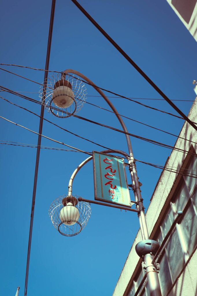 八王子の路地裏（どんどん横丁）にある、レトロなデザインの街灯を真下から見上げたカット。澄んだ青空と電線が背景に広がっている。