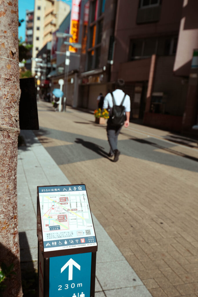 朝の八王子・ユーロード（西放射線ユーロード）。手前に街の案内看板が配置され、背景には通勤中の人物が歩いていく様子がボケて写っている。