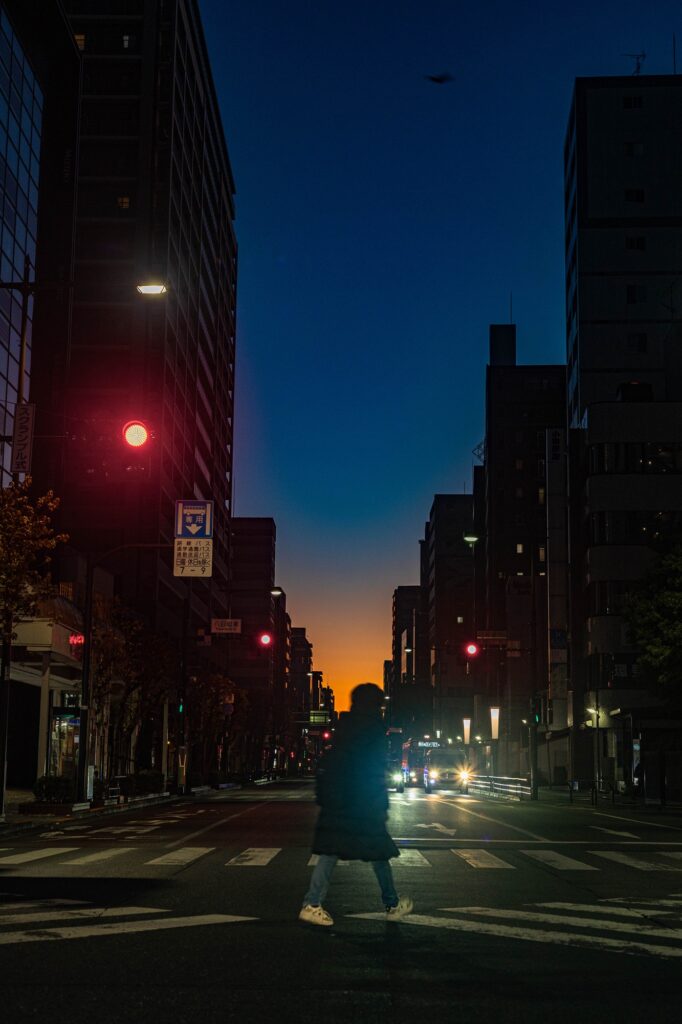 Home 早朝の八王子・八日町交差点。深い青色の空と鮮やかな朝焼けのグラデーションを背景に、横断歩道を渡る人のシルエットと街灯が輝く都会の風景。
