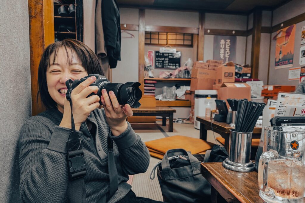 八王子、酒蔵「一平」。昭和の重低音とカメラ談義。 居酒屋の座敷で、キヤノンのミラーレスカメラを構えて満面の笑みを浮かべる女性のポートレート。