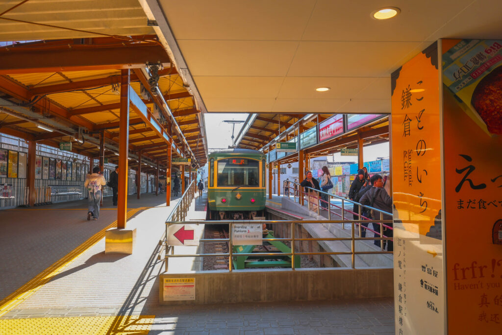 木造の屋根が特徴的な江ノ電鎌倉駅のホームに停車する、緑色の江ノ電車両と乗客の様子。