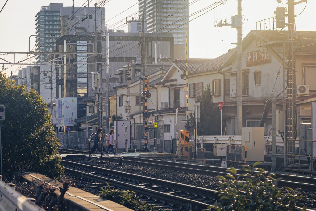 夕陽が差し込む八王子の踏切。黄金色の光の中で線路を渡る人々と、立ち並ぶ住宅街を捉えたエモーショナルな日常写真。
