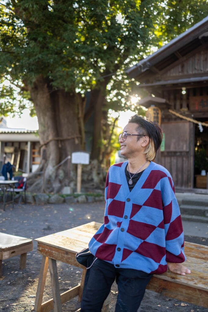 八王子いちょう祭りにて。大きな銀杏の木がある神社境内で、木漏れ日を浴びながら笑顔で木製のベンチに腰掛ける、青と赤のチェック柄カーディガンを着た男性のポートレート。