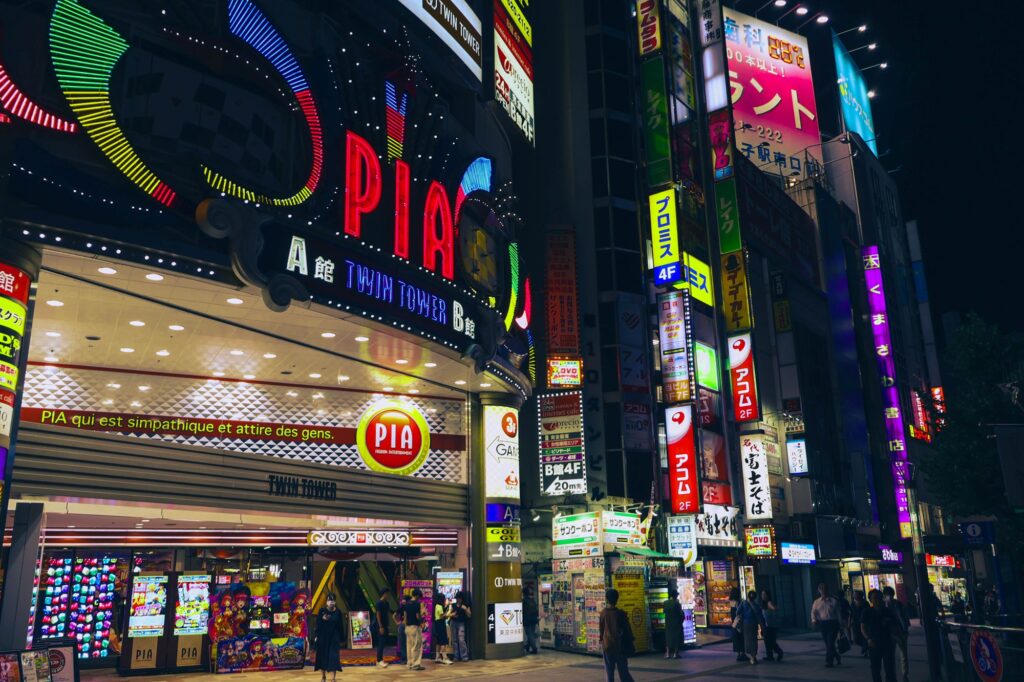 夜の八王子駅北口。パチンコ店「PIA」の巨大な赤いネオン看板を中心に、飲食店や消費者金融などの色とりどりの看板が密集し、夜空を明るく照らす繁華街の風景。