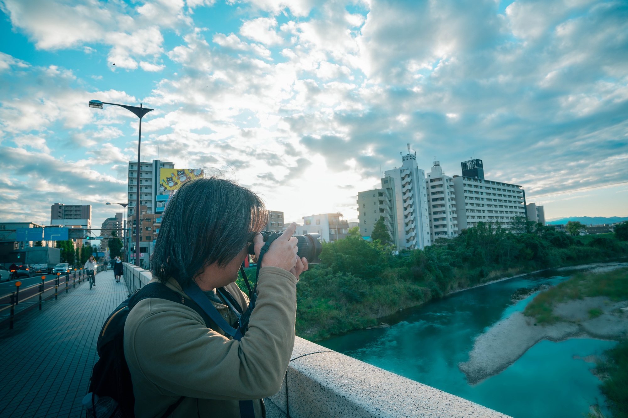 about 八王子の(浅川)に架かる大和田橋の上で、ドラマチックな雲と夕景に向かってカメラを構える澤井フォトグラファーの背中。