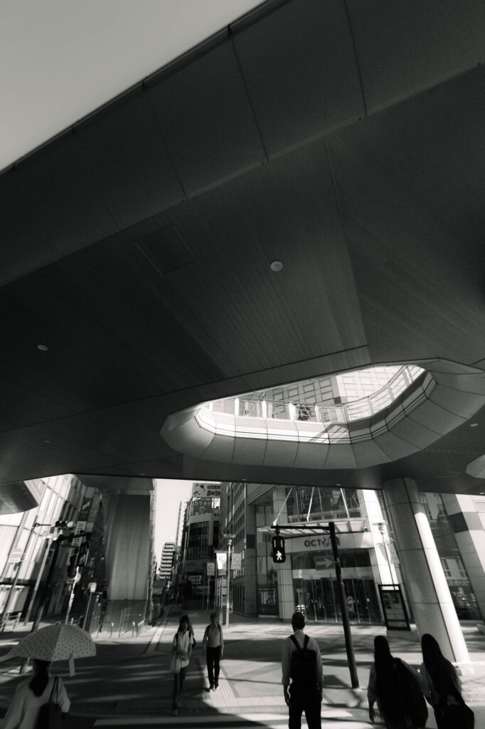 八王子駅北口ロータリーのマルベリーブリッジ下から撮影されたモノクロ写真。巨大な構造物の影と、楕円形の開口部から差し込む強い光が、歩行者たちのシルエットを強調している。