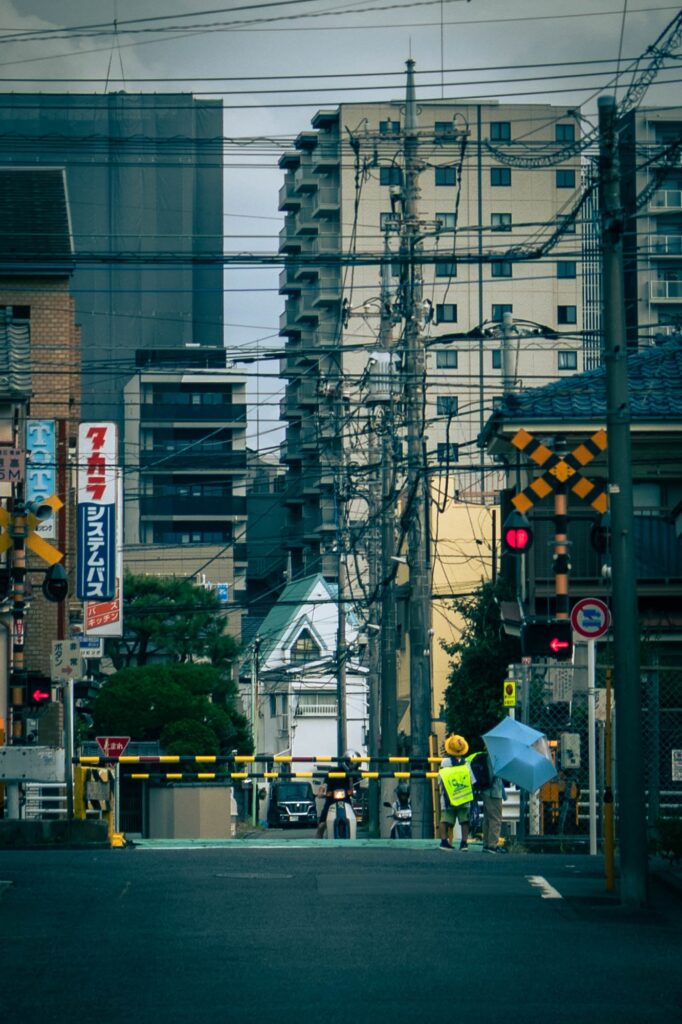 八王子市天神町付近の踏切風景。幾重にも重なる電線とビル群を背景に、遮断機の前に立つ黄色い帽子の小学生と傘をさした人の後ろ姿。