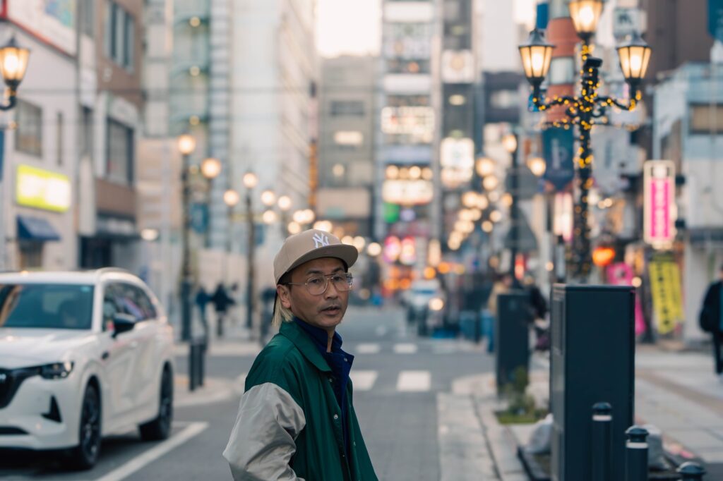 八王子のパーク一番街の通りで、緑のジャケットとキャップ姿で立つ男性のポートレート。