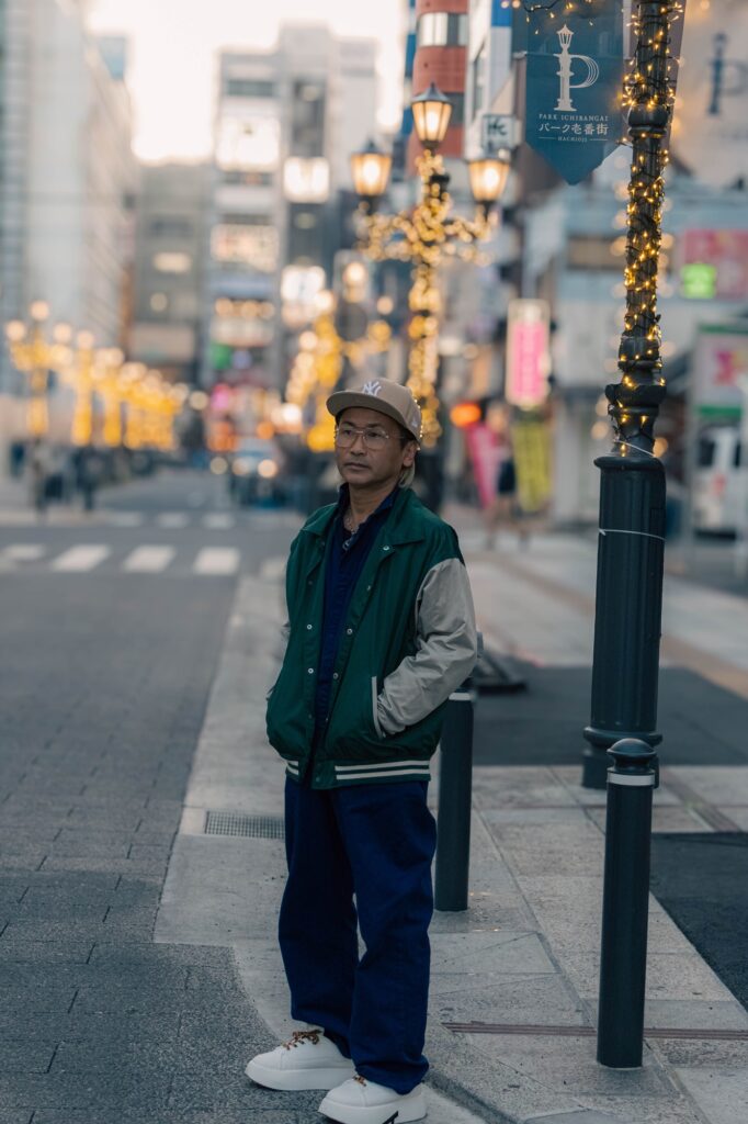 八王子、酒蔵「一平」。昭和の重低音とカメラ談義。 八王子のパーク一番街の通りで、緑のジャケットとキャップ姿で立つ男性のポートレート。背景にはイルミネーションの街灯がボケて写っている。