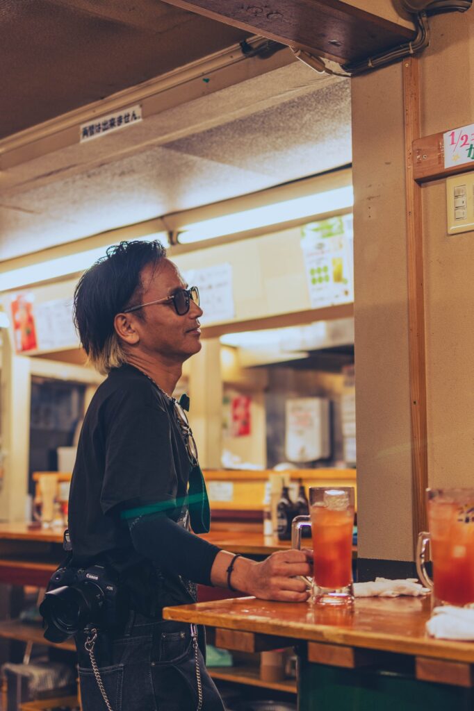 日野駅近くの立ち飲み屋。一眼レフカメラを腰に下げた男性が、カウンターでジョッキを手にリラックスしている横顔。温かみのある屋内の照明が印象的なスナップ。