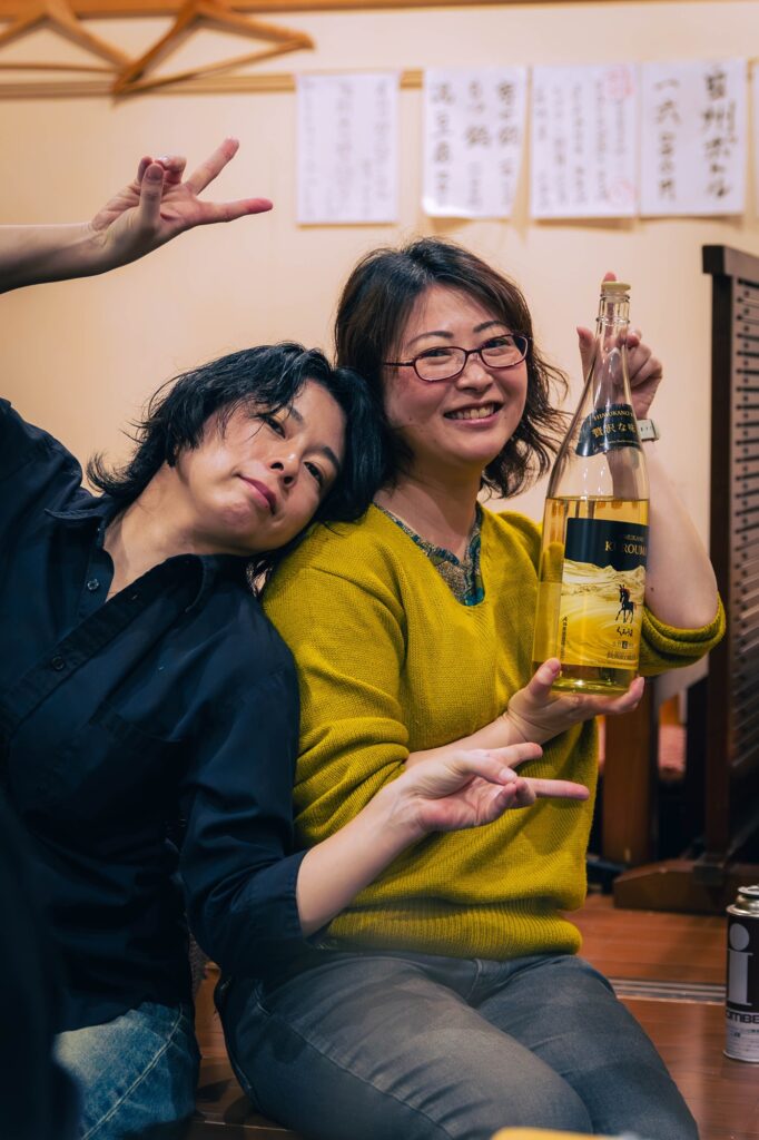 相模湖の静寂、西八王子の熱。仲間と囲む「痛風鍋」で締める、最高の休日。 西八王子「染井」にて、大きな酒瓶を手に笑顔でポーズをとる二人の女性。楽しげな宴会の雰囲気が伝わるポートレート。