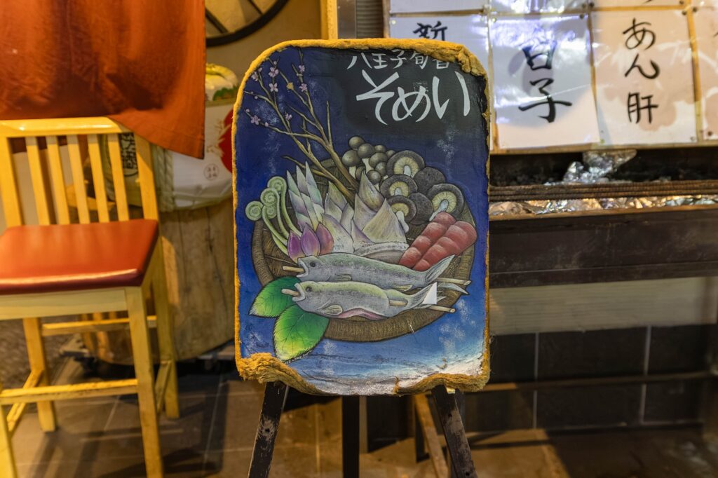 相模湖の静寂、西八王子の熱。仲間と囲む「痛風鍋」で締める、最高の休日。 西八王子「染井」の店先に置かれた、色鮮やかな料理のイラストが描かれた手書きの置き看板。