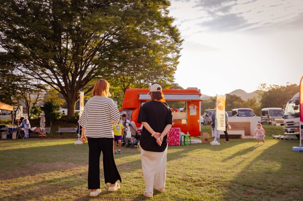 夕暮れ時の恩方マルシェ。大きな樹木とオレンジ色のキッチンカーを背景に、芝生の上に立つ人々と長く伸びる影、黄金色の西日が差し込む穏やかな風景。