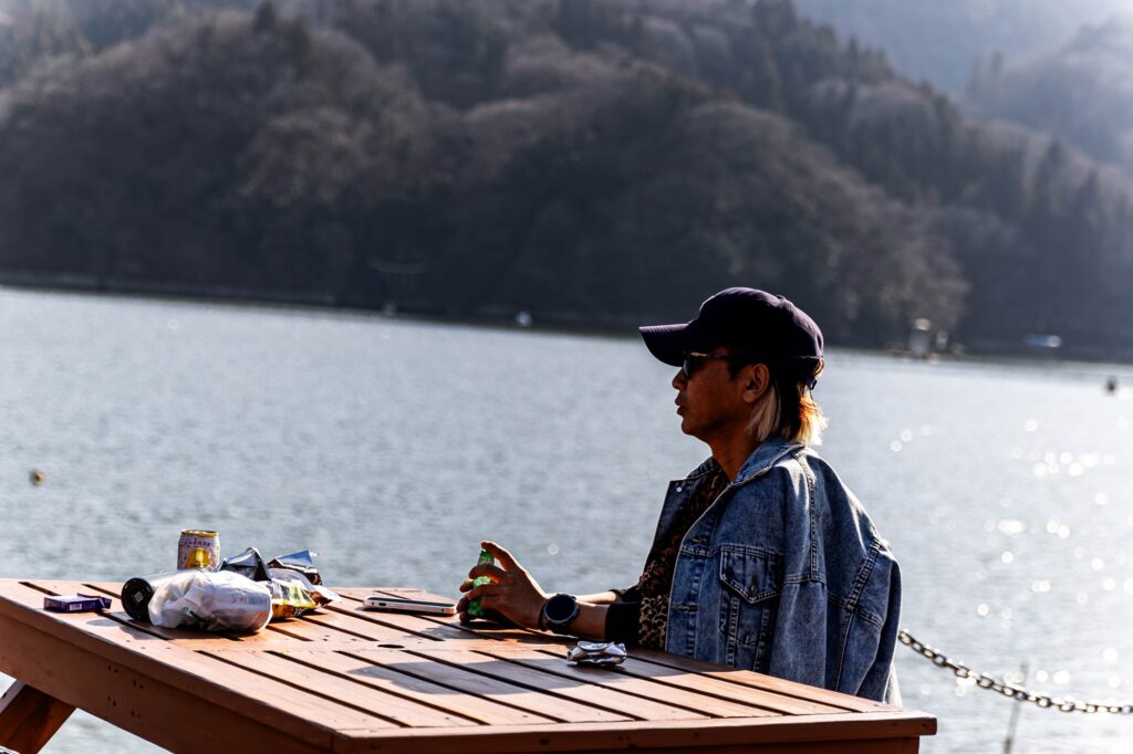 相模湖の静寂、西八王子の熱。仲間と囲む「痛風鍋」で締める、最高の休日。 陽光がきらめく湖を背景に、木製のテーブルでリラックスして座る男性の横顔。