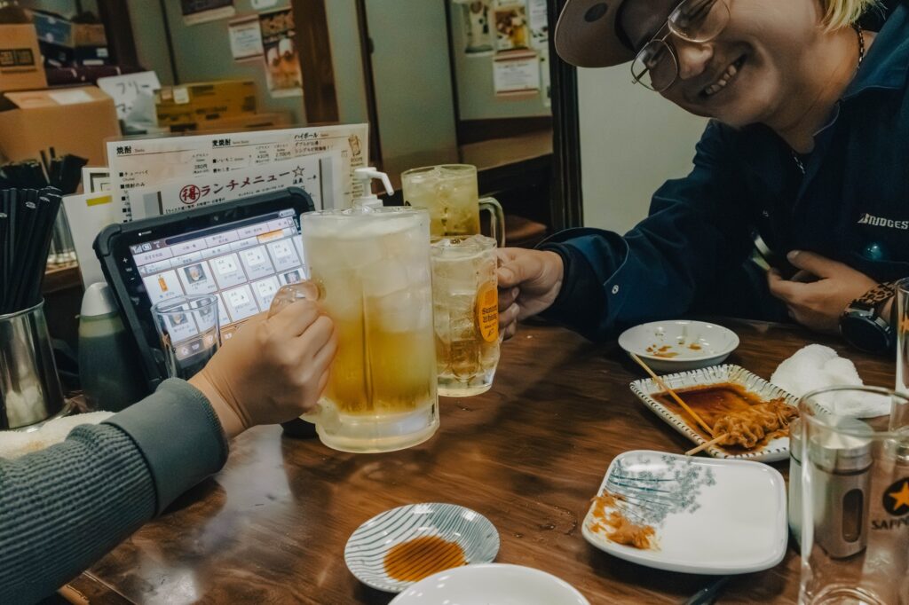 八王子、酒蔵「一平」。昭和の重低音とカメラ談義。 居酒屋のテーブルで、ビールジョッキとハイボールグラスを合わせて乾杯する様子。手前には注文用のタブレットが見える。