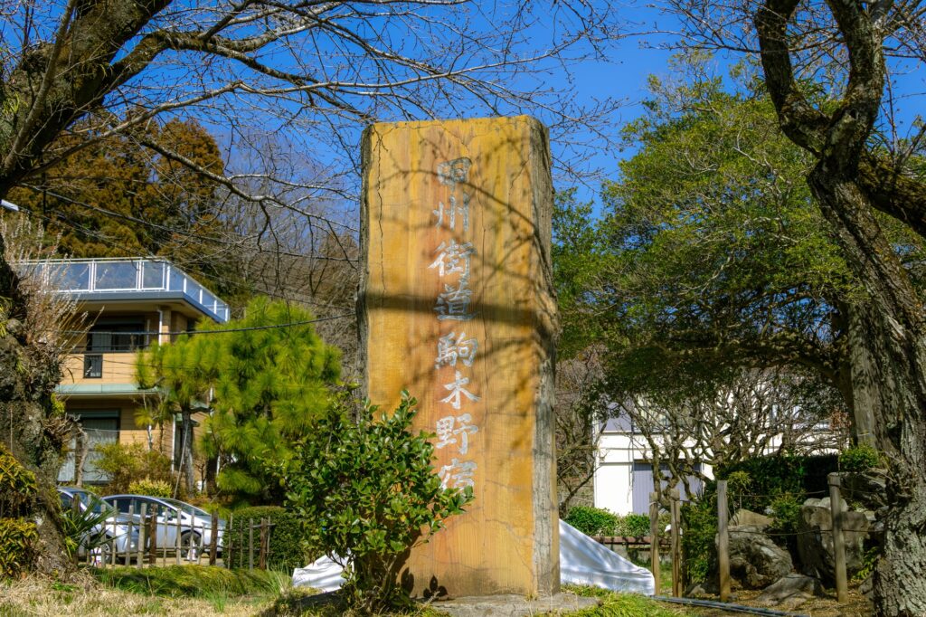 甲州街道・駒木野宿と刻まれた石碑。背後には青空と春の木々、石碑の表面には枝の影が美しく落ちている。